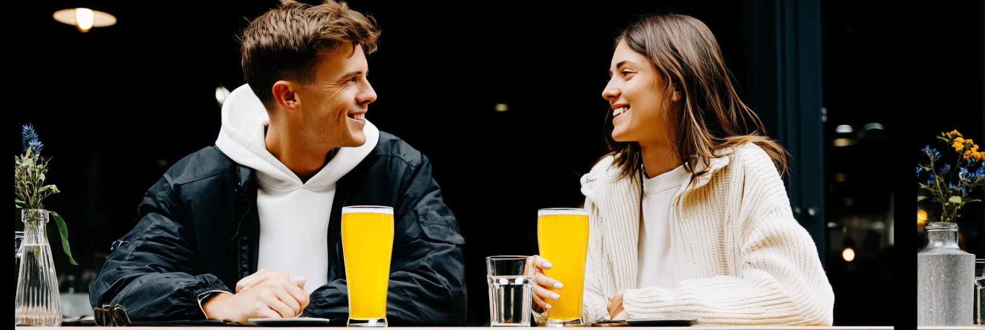 Um homem e uma mulher sentam-se frente a frente em uma mesa, sorrindo e conversando enquanto bebem cerveja em um bar moderno.